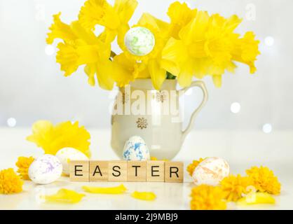Word Easter on wooden cubes. Easter composition of a bouquet of daffodils and multi-colored eggs against the background of blurry lights Stock Photo