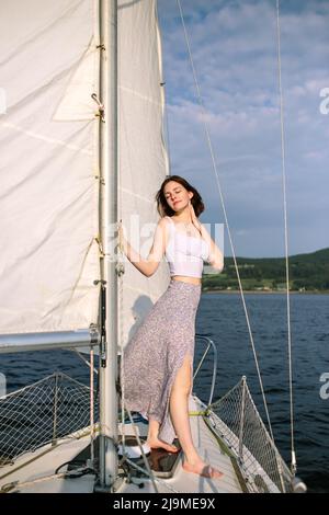 Full body of trendy young female tourist in stylish outfit standing on deck of modern yacht sailing in sea during summer holidays Stock Photo