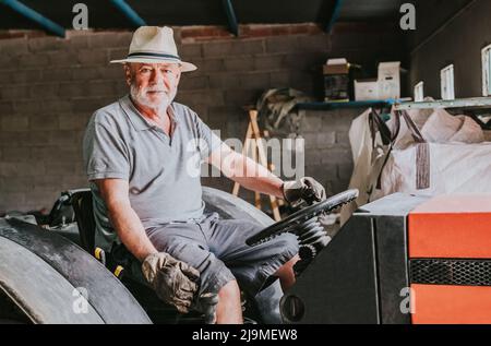 Senior male farmer in hat and gloves sitting on tractor parked in shabby garage in countryside and looking at camera Stock Photo