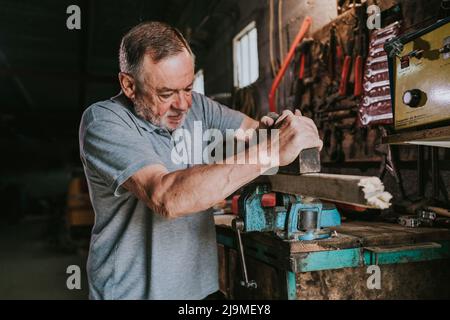 Focused mature male woodworker using jointer for planning wooden plank while working at shabby workbench in joinery workshop Stock Photo