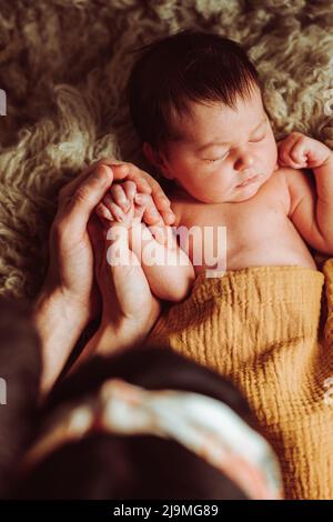 High angle of unrecognizable young mother and father with dark hair holding hand of adorable newborn baby lying and sleeping on soft bed in daylight Stock Photo