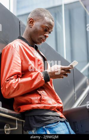 Side view of concentrated young African American guy in trendy red leather jacket messaging on mobile phone while leaning on wall on city street Stock Photo