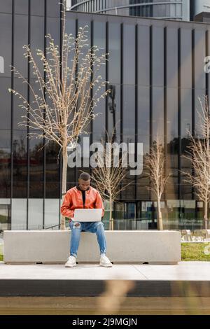 Focused young African American male freelancer in trendy outfit sitting looking at screen on stone bench and working remotely on laptop in university Stock Photo