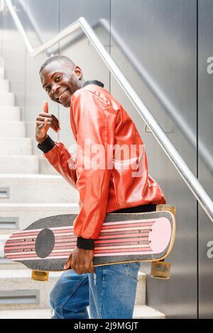 Side view of cheerful young African American guy in stylish outfit smiling at camera and showing call me gesture while walking upstairs with longboard Stock Photo