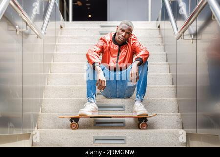 Young African American male in fashionable outfit sitting looking at camera on staircase with legs resting on longboard on city street Stock Photo