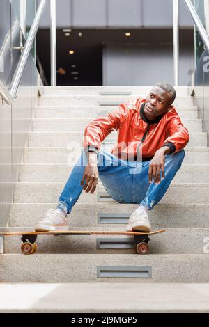 Young African American male in fashionable outfit sitting looking away on staircase with legs resting on longboard on city street Stock Photo