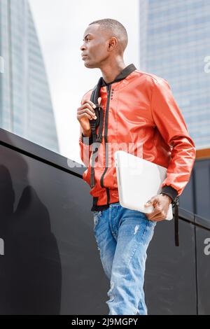 From below serious young trendy African American male in stylish outfit with backpack and laptop walking on street near contemporary building on sunny Stock Photo