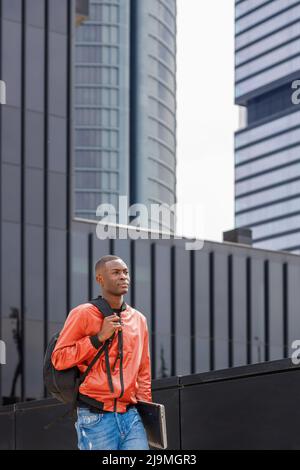 Serious young trendy African American male in stylish outfit with backpack and laptop walking on street near contemporary building on sunny day and lo Stock Photo