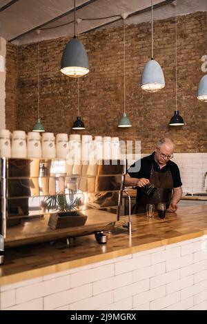 Concentrated mature male barista in apron pouring hot aromatic coffee from coffee maker into glass placed on wooden counter in cafe Stock Photo