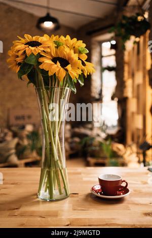 Glass vase with bunch of fresh sunflowers placed on wooden table near ceramic cup and saucer in cozy cafe Stock Photo