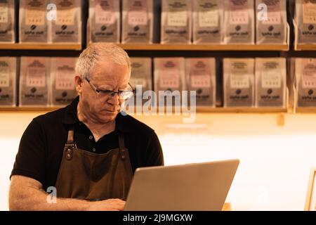 Concentrated mature male barista with gray hair in apron and eyeglasses using laptop while standing at counter in modern coffee shop Stock Photo