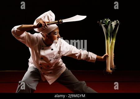 Funny young female clown in chef costume with makeup doing martial arts pose before cutting fresh leeks with knife on stage Stock Photo