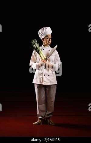 Full body of positive young female clown in chef outfit with makeup smiling and looking at camera while standing against black background with sharp k Stock Photo