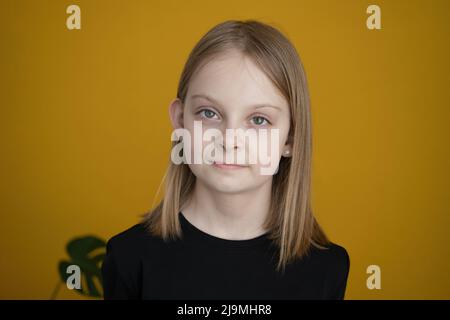 Adorable positive preteen girl with blond hair in black t shirt smiling and looking at camera against yellow background with green plants Stock Photo