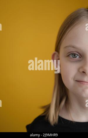 Half of face of adorable positive preteen girl with blond hair in black t shirt smiling and looking at camera against yellow background Stock Photo
