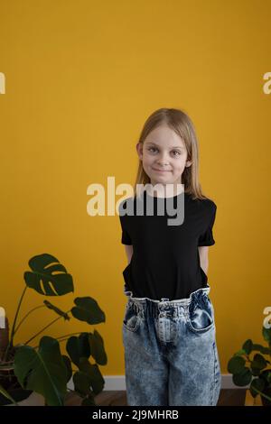 Adorable positive preteen girl with blond hair in black t shirt smiling and looking at camera against yellow background with green plants Stock Photo