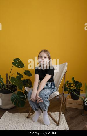 Adorable positive preteen girl with blond hair in black t shirt smiling and looking at camera relaxing on comfortable chair against yellow background Stock Photo