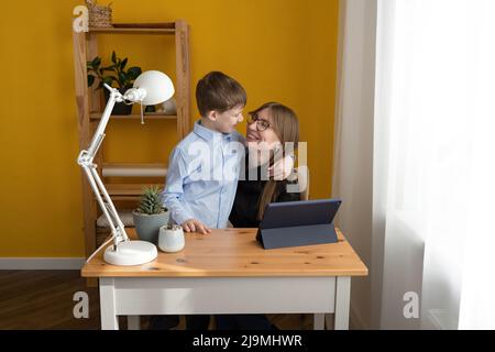 Child in blue shirt embracing happy mom in glasses sitting at table with tablet during remote work from home Stock Photo