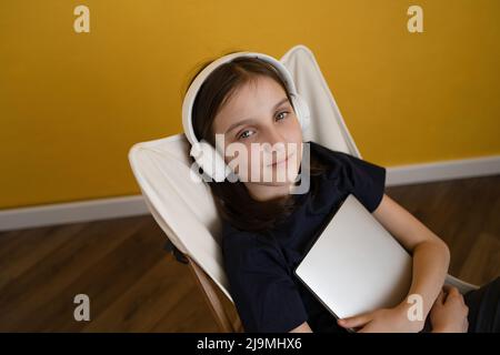 From above of calm preteen girl with laptop in hands in casual clothes relaxing on chair and listening to music in headphones at home Stock Photo