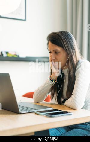 Thoughtful young Hispanic female freelancer with long dark hair in casual clothes and earphones sitting at wooden table with hand on cheek and working Stock Photo