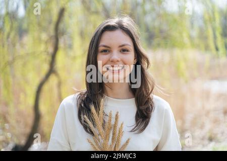 Young Woman with long dark hair and brown eyes holding bunch of wheat spikelets and looking at camera in countryside Stock Photo