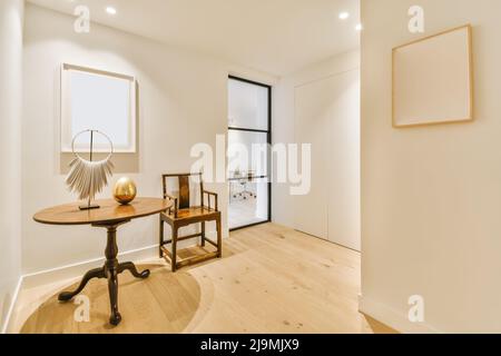 Round retro table with golden egg and unusual decoration located near wooden chair in corridor with white walls and empty frames Stock Photo