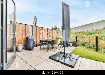 Folded parasol and rubbish bin placed on tiled floor near table and chairs against wooden fence and blue sky outside house Stock Photo