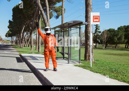 Full body of unrecognizable male cosmonaut in orange suit and helmet catching bus while standing on sidewalk against cloudless blue sky in city Stock Photo
