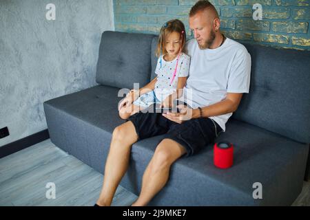 Father and daughter testing talking smart wireless speaker, controlling home devices with a voice commands, sitting on sofa at home, Concept of smart Stock Photo