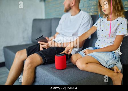 Father and daughter testing talking smart wireless speaker, controlling home devices with a voice commands, sitting on sofa at home, Concept of smart Stock Photo