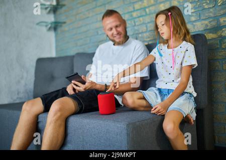 Father and daughter testing talking smart wireless speaker, controlling home devices with a voice commands, sitting on sofa at home, Concept of smart Stock Photo