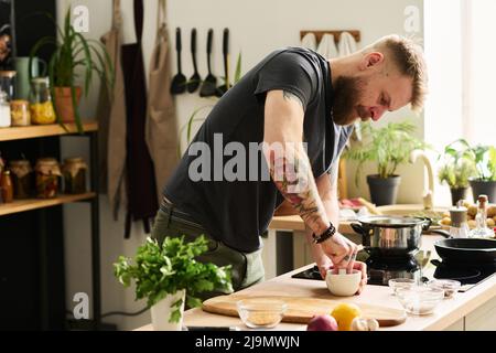 Handsome young Caucasian man with tattoos on arms standing in kitchen crushing spices with mortar and pestle Stock Photo