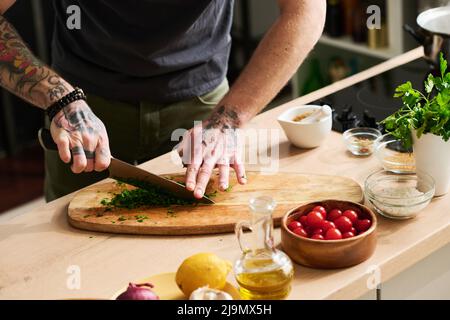 Horizontal medium section shot of unrecognizable chef with tattooes on arms chopping fresh cilantro or parsley on cutting board Stock Photo