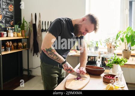 Young adult tattooed Caucasian man with beard and moustache on face standing in kitchen cutting red onion on wooden board Stock Photo
