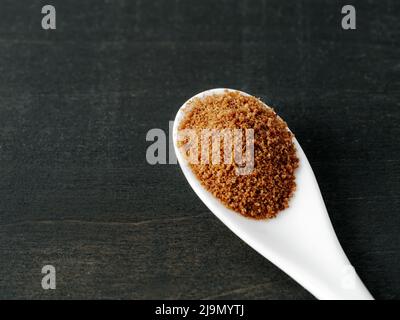 Spoon of coconut brown sugar on dark wooden background. Top view of full white ceramic spoon with raw organic coconut sugar from crystallized sap of cut flower buds coconut palm. Copy space. Closeup Stock Photo