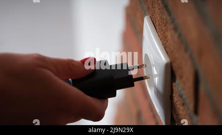 Adult woman holds in hands and connects black electric plug to socket at home Stock Photo