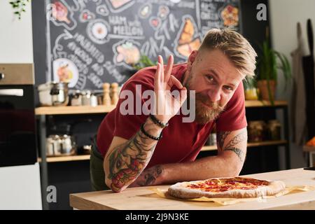 Joyful Caucasian man with veard on face posing on camera with freshly baked pizza showing OK hand gesture Stock Photo