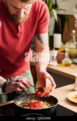 Vertical shot of bearded man wearing casual clothes cooking meal in kitchen at home adding tomato sauce to ingredients in frying pan Stock Photo