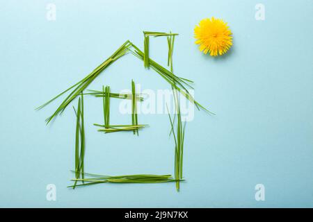 house made of grass bades with chimney and a dandelion as the sun. Grass straws on light blue background Stock Photo