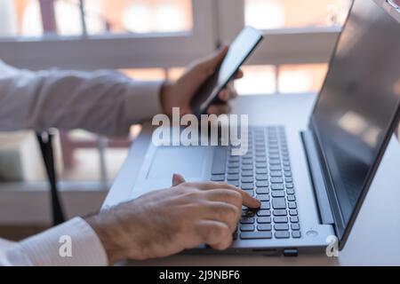 Man typing on a laptop and checking the mobile phone while teleworking. Stock Photo