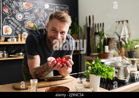 Cheerful young Caucasian man with beard on face and tattoos on arms holding cherry tomatoes in hands smiling at camera Stock Photo