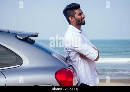 happy indian man in white shirt collar buying new car and showing the key, sitting in car on beach sea india octan Goa . a trip to the beach in car Stock Photo