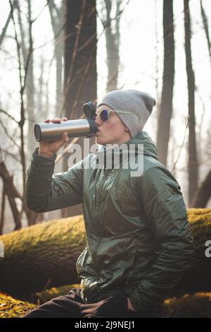 experienced hiker sitting on a tree trunk in the wilderness, drinking his hot tea from a thermos during a break in the walk. Real people discovering t Stock Photo