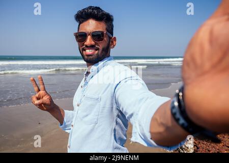 Young african american man in blue shirt and jeans making a selfie portrait on the front camera of a smartphone on the ocean beach. Stock Photo