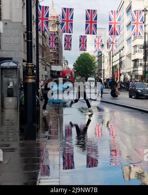 Mans leaps across the puddles of water in Oxford Street after heavy rain, with Union Jack flags reflected in the water. Stock Photo