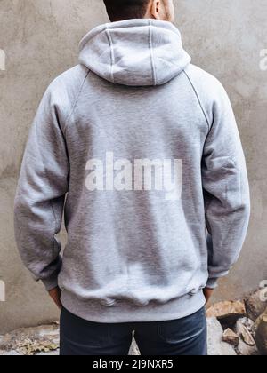 Back view of young man in a hoodie with hood for advertising with ability to apply logo. A man stands on a background of light gray concrete wall. Des Stock Photo