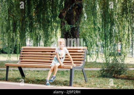 A cute boy is sitting on a bench in the park under a big green tree and resting. Stock Photo