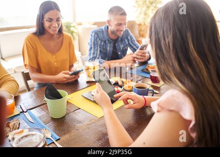 Group of people using phone smiling in a cafe bar. Friends having breakfast and watching mobile. Stock Photo