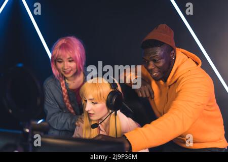 young Caucasian female player plays video games, Asian woman and Afro-American watch the game copy space dark background . High quality photo Stock Photo