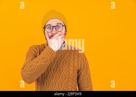 Shocked devastated caucasian hipster man in his 30s in a knitted sweater and matching beanie covering his beard with one hand over yellow background. High quality photo Stock Photo